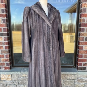 May include: A full-length, gray fur coat with a wide collar and long sleeves. The coat is displayed on a mannequin in front of a window. The text "CHICKASAW FURS" and a logo are visible in the background.