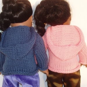 Könnte beinhalten: Zwei Puppen tragen Strickpullover mit Kapuze. Eine Puppe trägt einen marineblauen Pullover und eine lila Hose. Die andere Puppe trägt einen rosa Pullover und eine braune Hose. Die Puppen sind dem Betrachter abgewandt.
