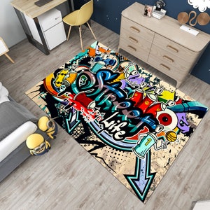 Puede incluir: Una alfombra rectangular con un vibrante dise&ntilde;o de graffiti. La alfombra presenta colores llamativos, incluyendo rojo, azul, amarillo y morado, con la palabra "Life" prominentemente mostrada. El dise&ntilde;o incluye botes de pintura en aerosol, flechas y otros elementos de arte urbano.