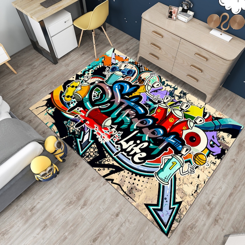 Puede incluir: Una alfombra rectangular con un vibrante dise&ntilde;o de graffiti. La alfombra presenta colores llamativos, incluyendo rojo, azul, amarillo y morado, con la palabra "Life" prominentemente mostrada. El dise&ntilde;o incluye botes de pintura en aerosol, flechas y otros elementos de arte urbano.