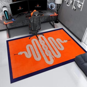 Könnte beinhalten: Ein rechteckiger, orangefarbener Teppich mit marineblauem Rand und einem großen, weiß-braunen Schlangenmuster. Der Teppich befindet sich in einem Raum mit Gaming-Setup, einschließlich Schreibtisch, Stuhl und zwei Monitoren.