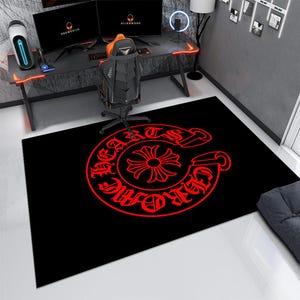 May include: A rectangular area rug featuring a black background with a large, circular red graphic in the center. The design includes a stylized cross symbol surrounded by text that reads CHROME HEARTS in a bold, gothic-inspired typeface.