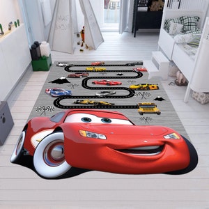 May include: A red cartoon race car with a smiling face is featured on a gray and white rug with a black race track design. The rug also features smaller yellow and blue race cars, a yellow school bus, and black and white checkered flags.