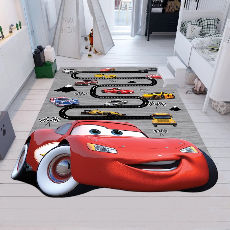 May include: A red cartoon race car with a smiling face is featured on a gray and white rug with a black race track design. The rug also features smaller yellow and blue race cars, a yellow school bus, and black and white checkered flags.