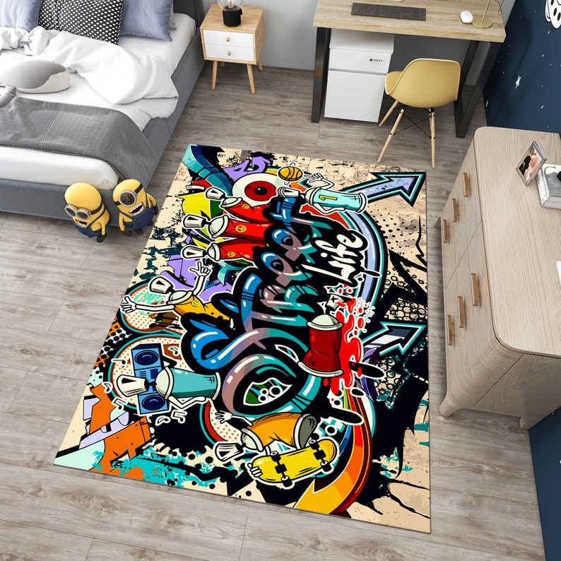 Puede incluir: Una alfombra rectangular con un vibrante dise&ntilde;o de graffiti. La alfombra presenta colores llamativos, incluyendo azul, rojo, amarillo y negro, con botes de pintura en aerosol, flechas y letras estilizadas. El dise&ntilde;o est&aacute; sobre un fondo beige.