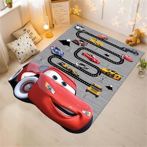May include: A red cartoon race car with a smiling face is on a gray rug with a black race track design. The rug features other cartoon cars, including a yellow school bus, a black race car, and a blue car.