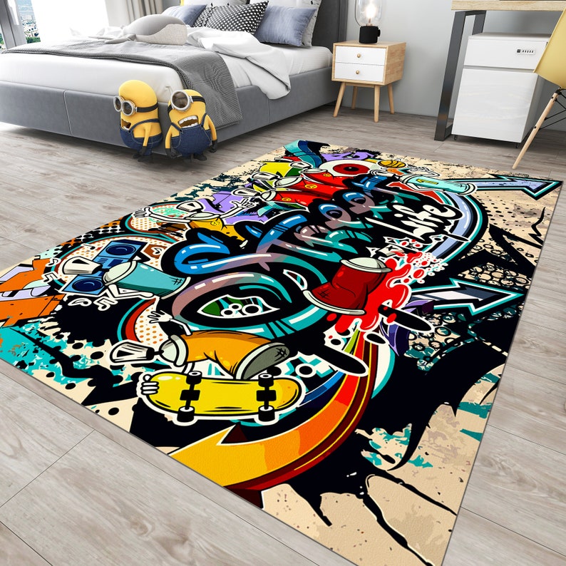 Puede incluir: Una alfombra rectangular con un vibrante dise&ntilde;o de graffiti. La alfombra presenta un monopat&iacute;n amarillo, botes de pintura en aerosol y letras en negrita en una mezcla de colores que incluyen rojo, azul y amarillo. El fondo es una mezcla de negro, beige y blanco.