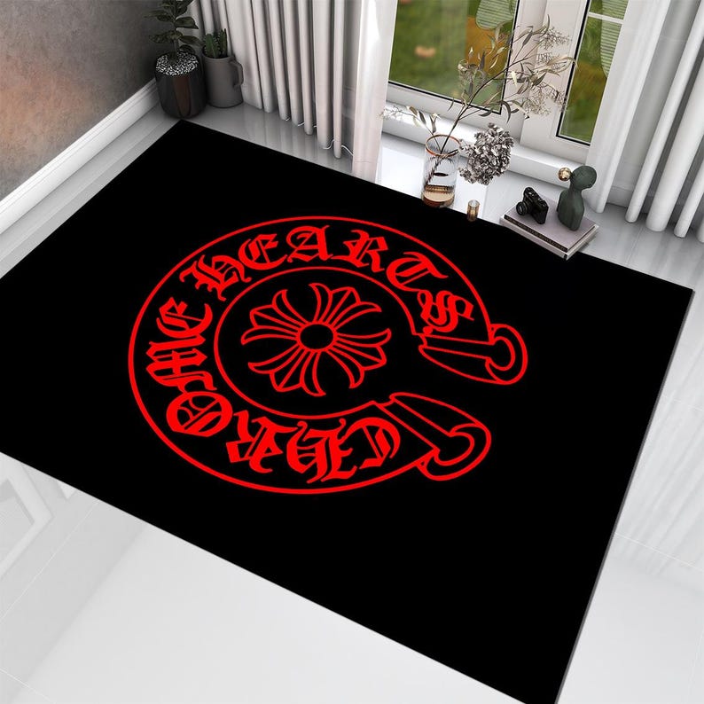 May include: A rectangular black area rug featuring a large, centered red graphic. The design consists of a circular emblem with a stylized cross in the middle, surrounded by the text "CHROME HEARTS" written in a bold, gothic-style typeface.