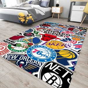 May include: A colorful rug with a white background featuring various NBA team logos, including the Chicago Bulls, Los Angeles Lakers, Philadelphia 76ers, and New Orleans Pelicans.