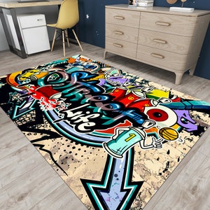 Puede incluir: Una alfombra rectangular con un dise&ntilde;o de grafiti audaz. La alfombra presenta una flecha grande, botes de pintura en aerosol y las palabras "Live Life" en un estilo de arte urbano colorido. La paleta de colores incluye negro, azul, rojo, amarillo y blanco.
