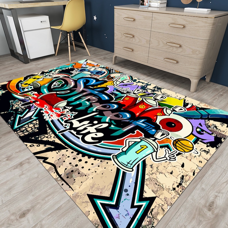 Puede incluir: Una alfombra rectangular con un dise&ntilde;o de grafiti audaz. La alfombra presenta una flecha grande, botes de pintura en aerosol y las palabras "Live Life" en un estilo de arte urbano colorido. La paleta de colores incluye negro, azul, rojo, amarillo y blanco.