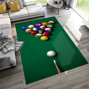 May include: A green rectangular rug designed to resemble a pool table, complete with a rack of billiard balls in various colors and a white cue ball. A brown cue stick rests near the cue ball. The rug is placed on a light wood floor.