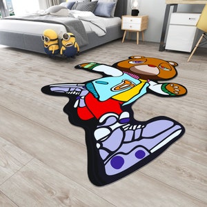 May include: A black and white rug with a cartoon bear wearing a blue, yellow, and red outfit. The bear is lying on its back with its arms and legs outstretched. The rug has a black outline and is on a wooden floor.