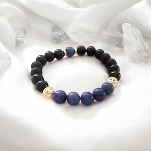 Può includere: Un braccialetto con perline nere e blu con accenti dorati. Il braccialetto è realizzato con perline di roccia lavica nera e perline di lapislazzuli blu.