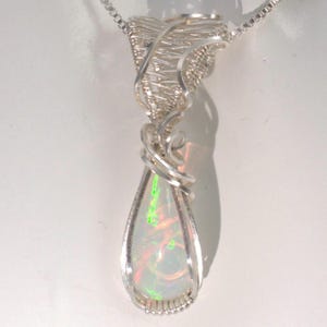 Può includere: Collana con pendente in argento fatta a mano con un'opale a forma di pera. L'opale mostra colori iridescenti verdi, rossi e bianchi. La gemma è incastonata in un'impostazione avvolta in filo metallico, sospesa da una delicata catena.