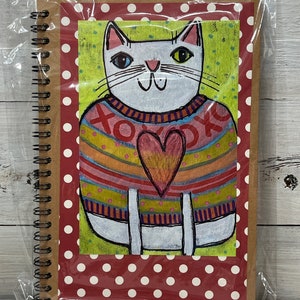May include: A white cat wearing a colorful sweater with a heart on the front. The cat is on a red background with white polka dots. The sweater has the text "XOXO" on it.