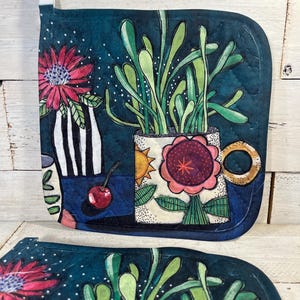 May include: A quilted, square potholder with a dark teal background features a floral design. A large flower in a mug and a vase with a red flower are depicted. A white loop is attached for hanging.