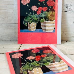 May include: Greeting cards featuring a floral illustration of geraniums in pots. The artwork depicts pink and red flowers with green leaves against a dark background. The pots have a polka dot and striped design, framed by a pink border.
