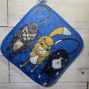 May include: Blue quilted pot holder featuring three cartoon cats in gray, yellow, and black. The cats are depicted with smiling faces and long tails. The pot holder has a hanging loop.