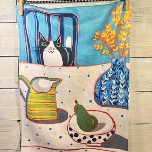 May include: A decorative kitchen towel featuring a whimsical illustration. The design includes a black and white cat, a yellow and green pitcher, a green pear in a bowl, and a blue vase with yellow flowers. The towel is hanging from a wooden clothespin.