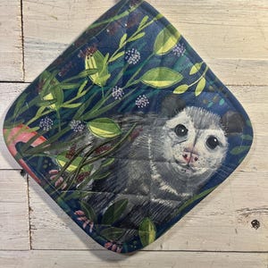 May include: A quilted, diamond-shaped potholder featuring a detailed illustration of an opossum amidst lush greenery. The design includes a dark blue background with floral and leaf patterns, and a white hanging loop.