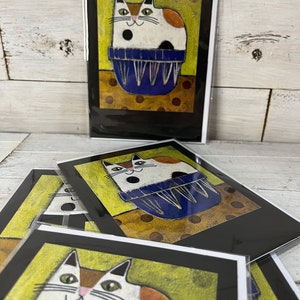 May include: A set of greeting cards featuring a white cat with orange and black markings sitting in a blue bowl. The cat has a black spot on its belly and green eyes. The background is yellow with brown polka dots.