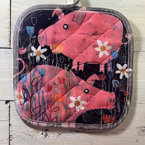 May include: Square quilted pot holder featuring two pink pigs with black eyes and white flower accents, set against a dark blue floral background. The pot holder has a gray border and a white fabric loop for hanging.