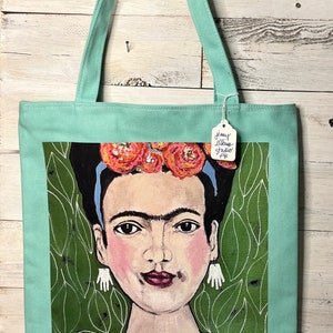 May include: A turquoise tote bag with a green background featuring a painting of Frida Kahlo. The painting shows Frida with a flower crown, blue headband, and a blue necklace. The text "Jenny Ellen Studio FB" is on a white tag attached to the bag.