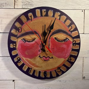 May include: A round wall clock with a painted sun face design. The clock face is yellow with a smiling face, closed eyes, and red cheeks. The clock has black hands and a red second hand. The outer edge is purple with white numbers.