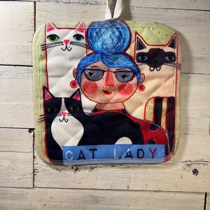 May include: A square, quilted pot holder with a whimsical design. It features a woman with blue hair and glasses surrounded by three cats. The words "CAT LADY" are printed on a blue ribbon at the bottom. The pot holder has a loop for hanging.