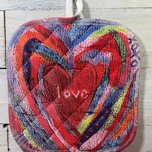 May include: A quilted, square-shaped potholder with a heart design. The heart is red with the word "love" embroidered in white. The potholder features a colorful, abstract pattern with a white loop for hanging.