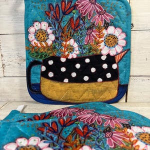 May include: Two turquoise pot holders with a floral design. One pot holder features a black polka-dotted pitcher filled with white and pink flowers. The other pot holder is partially visible, showing a similar floral pattern. Both have a loop for hanging.