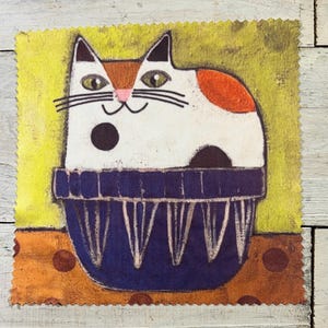 May include: A whimsical illustration of a white cat with orange and black markings, sitting in a blue bowl. The cat has green eyes and a pink nose. The background is yellow with an orange and brown polka dot pattern.