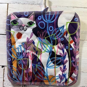 May include: Square quilted pot holder featuring a whimsical cat design with green eyes, a red spot, and a smiling face. The design includes colorful floral elements and a purple border. Includes a white loop for hanging.