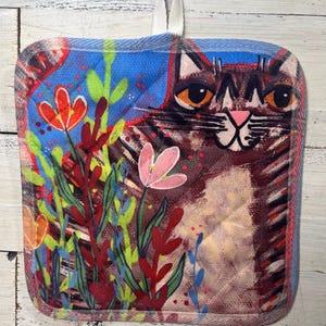 May include: Square quilted pot holder featuring a whimsical cat design. The cat has large orange eyes and a pink nose, surrounded by colorful flowers and foliage. The pot holder has a blue border and a white hanging loop.