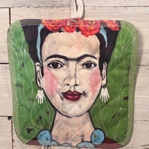 May include: Square quilted pot holder featuring a portrait of Frida Kahlo. The artwork includes a floral headdress, red lips, and green leaf background. The pot holder has a green border and a white loop for hanging.