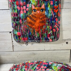 May include: A square, quilted pot holder featuring a vibrant, colorful design. The artwork depicts an orange tabby cat amidst a field of flowers in shades of pink, red, blue, and green. A white loop is attached for hanging.
