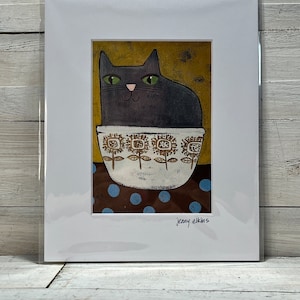 May include: A framed art print featuring a stylized black cat with green eyes, sitting in a white bowl with a floral design. The background is gold, and the base has a brown polka dot pattern. The artist's signature, "jenny elkins", is in the bottom right corner.