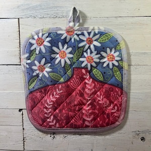 May include: Square quilted pot holder with a floral design. The top half features white daisies on a blue background, while the bottom half has a red vase design. The pot holder has a hanging loop.
