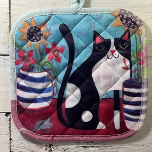 May include: Quilted pot holder featuring a black and white cat with green eyes, sitting in front of a blue floral background. The pot holder has a loop for hanging and a red border. Decorative vases with flowers and a mouse are also visible.