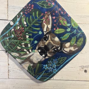 May include: A quilted, diamond-shaped potholder featuring a deer's face surrounded by green leaves, red berries, and blue flowers against a dark blue background. The potholder has a white loop for hanging and is likely used for kitchen or cooking purposes.