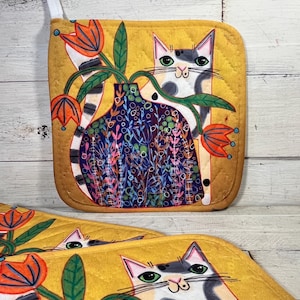 May include: Yellow quilted pot holders with a cat and floral design. The cat is white, gray, and black with green eyes. The flowers are orange and the vase is blue with colorful floral patterns. The pot holders have a loop for hanging.