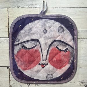 May include: Square quilted pot holder featuring a whimsical moon face design with closed eyes and rosy cheeks. The pot holder has a purple border and a loop for hanging. The moon face is painted in shades of gray, white, and red.