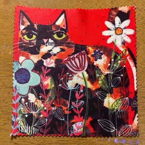 May include: A square fabric with a colorful cat design. The cat has orange, black, and white fur with green eyes, set against a red background. The fabric is decorated with various flowers in shades of blue, pink, and white.