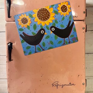 May include: A vintage pink refrigerator with a decorative magnet. The magnet features two black birds and sunflowers on a blue background. The refrigerator has a handle and the word "Refrigerator" is written on the front.