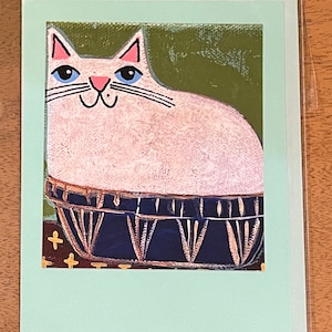May include: A white cat with blue eyes sits in a blue and gold patterned bowl. The cat has pink ears and a pink nose. The bowl is on a brown and white patterned background.