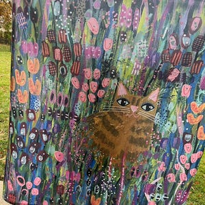 May include: A colorful tapestry featuring a brown tabby cat sitting in a field of flowers. The flowers are painted in shades of pink, purple, blue, green, and orange. The cat has green eyes and a white patch on its chest.