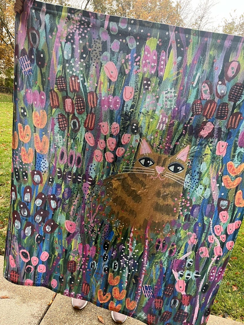 May include: A colorful tapestry featuring a brown tabby cat sitting in a field of flowers. The flowers are painted in shades of pink, purple, blue, green, and orange. The cat has green eyes and a white patch on its chest.