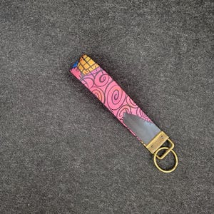 May include: A pink and black patterned key chain with a gold metal key ring. The key chain has a swirl design.