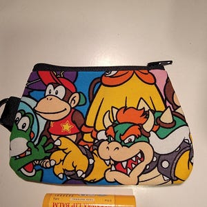 May include: A colorful zippered pouch featuring cartoon characters, including a monkey, a turtle, and a dragon. The pouch is rectangular with a black zipper and strap. A tube of Burt's Bees lip balm is next to the pouch.
